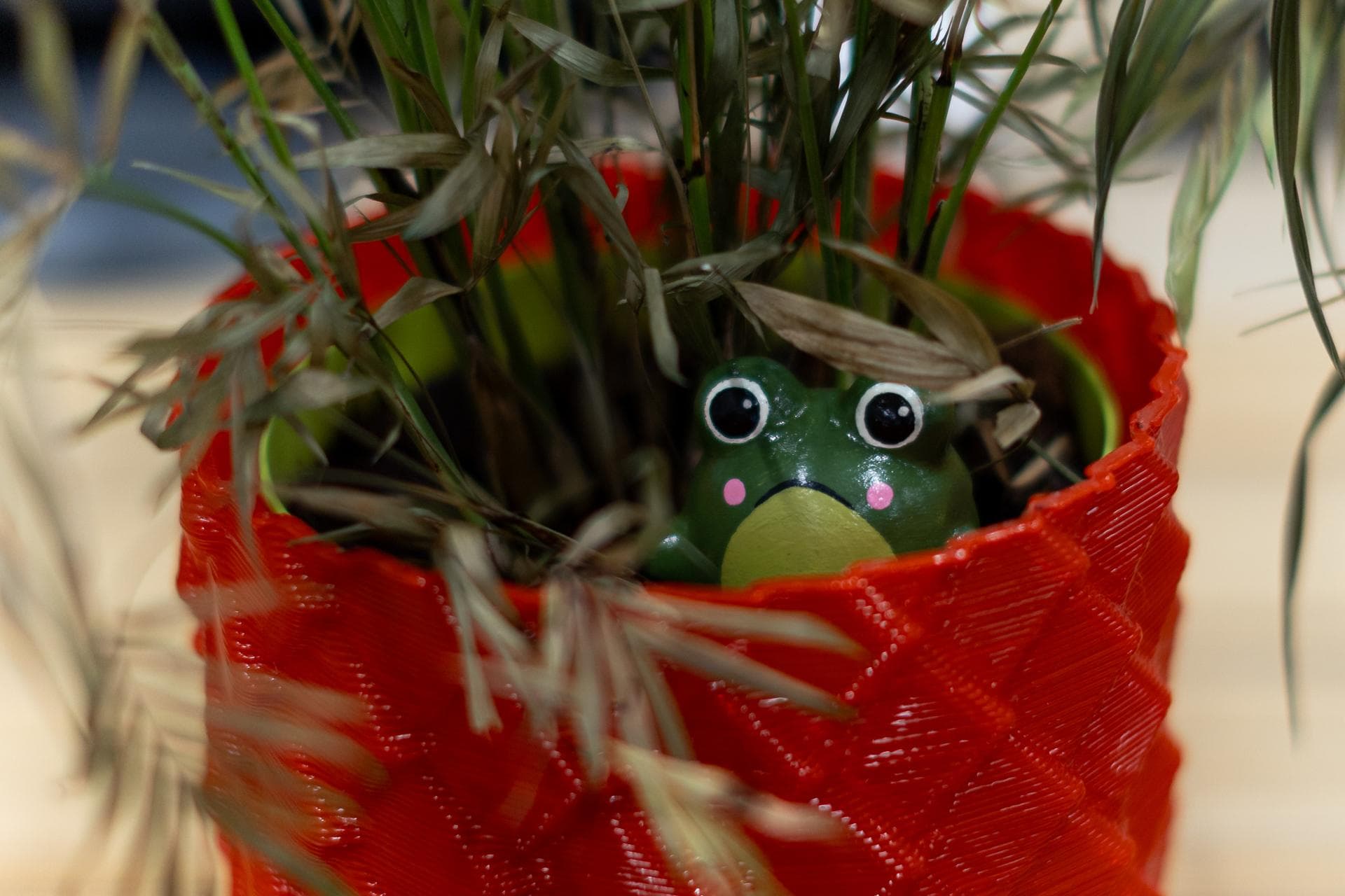 A hand-painted green frog peeking out of a red 3D-printed plant pot, half hidden by dry grass.
