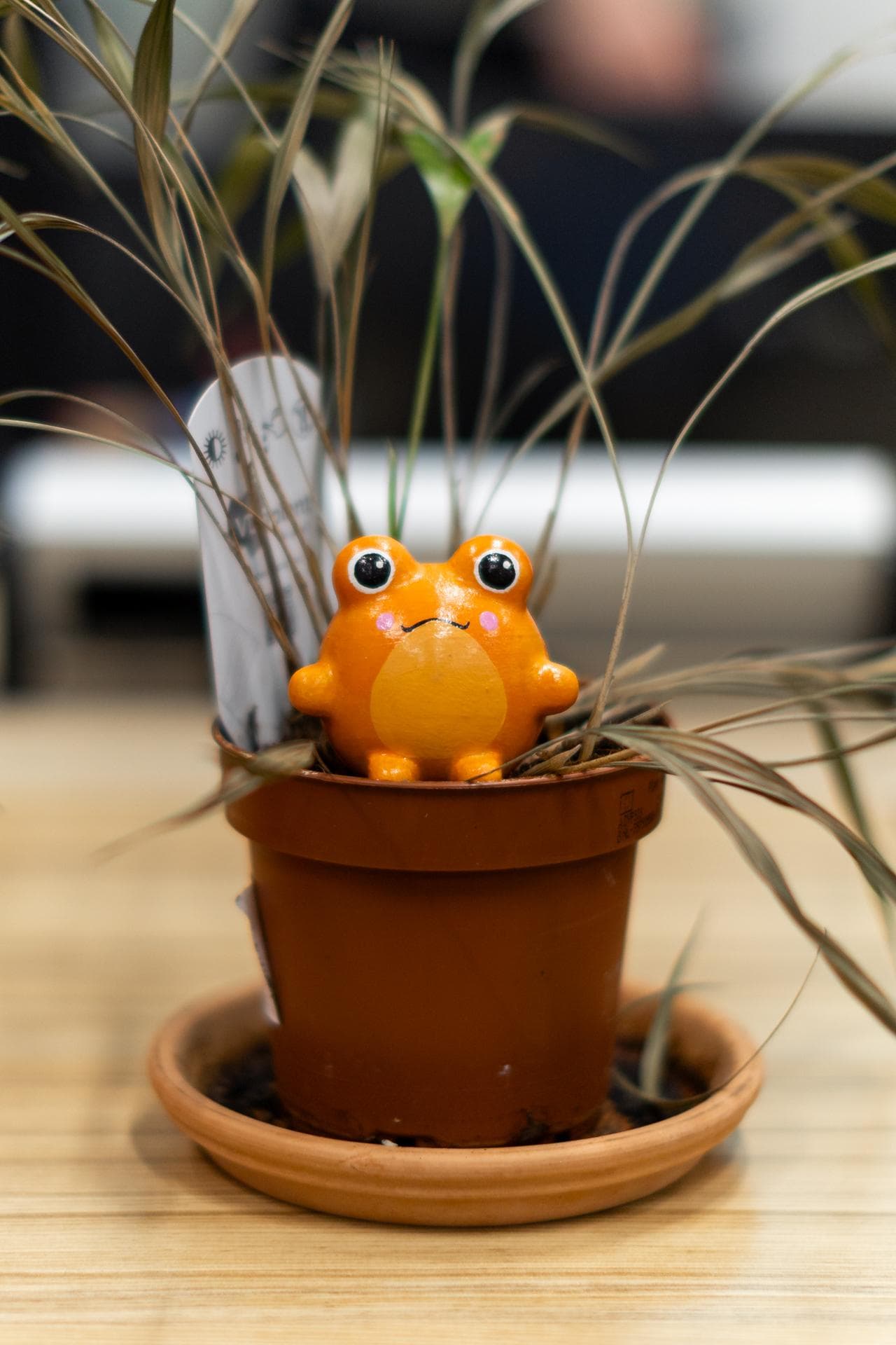 An orange Sprooty frog sitting in a terracotta pot of dry ornamental grass.