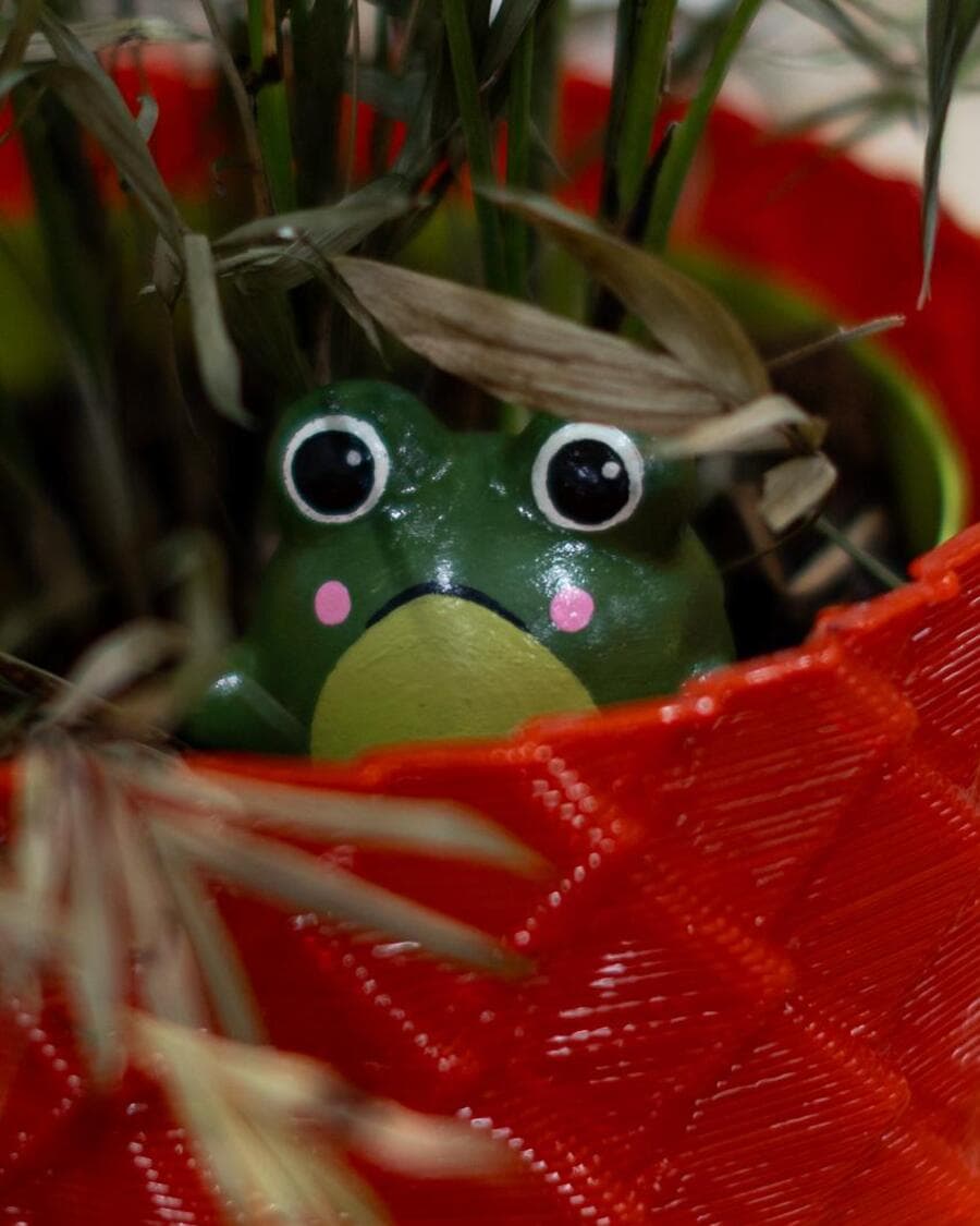 A green Sprooty frog peering over the rim of a red 3D-printed pot.