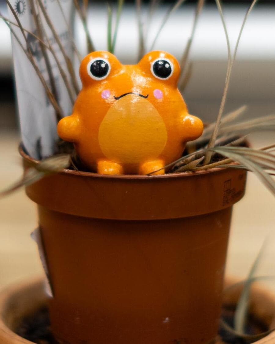 An orange Sprooty frog sitting in a terracotta pot of dry ornamental grass.